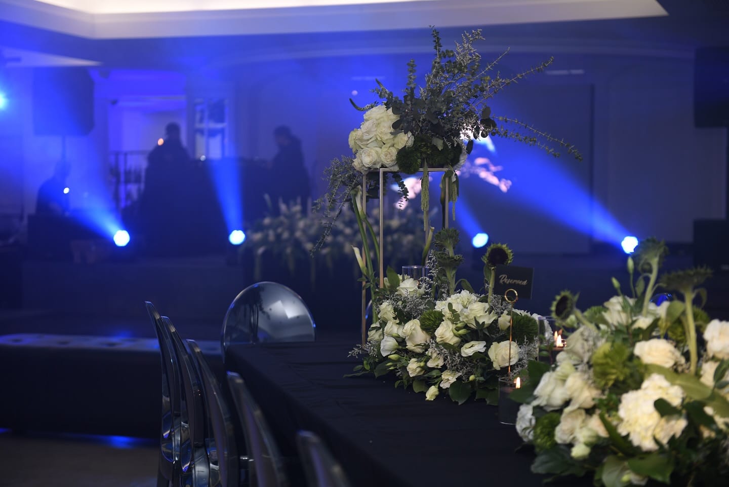 Gold stand of white roses and trailing eucalyptus beside low white centerpieces and a Reserved sign on a black table, with silhouetted guests and blue haze in the ballroom behind.