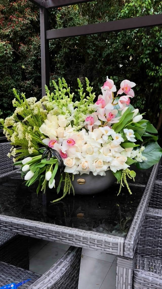 White roses, white tulips, pale pink cymbidium orchids, white snapdragons, stock, monstera and trailing green amaranthus in a grey concrete bowl on a black glass patio table under a pergola.
