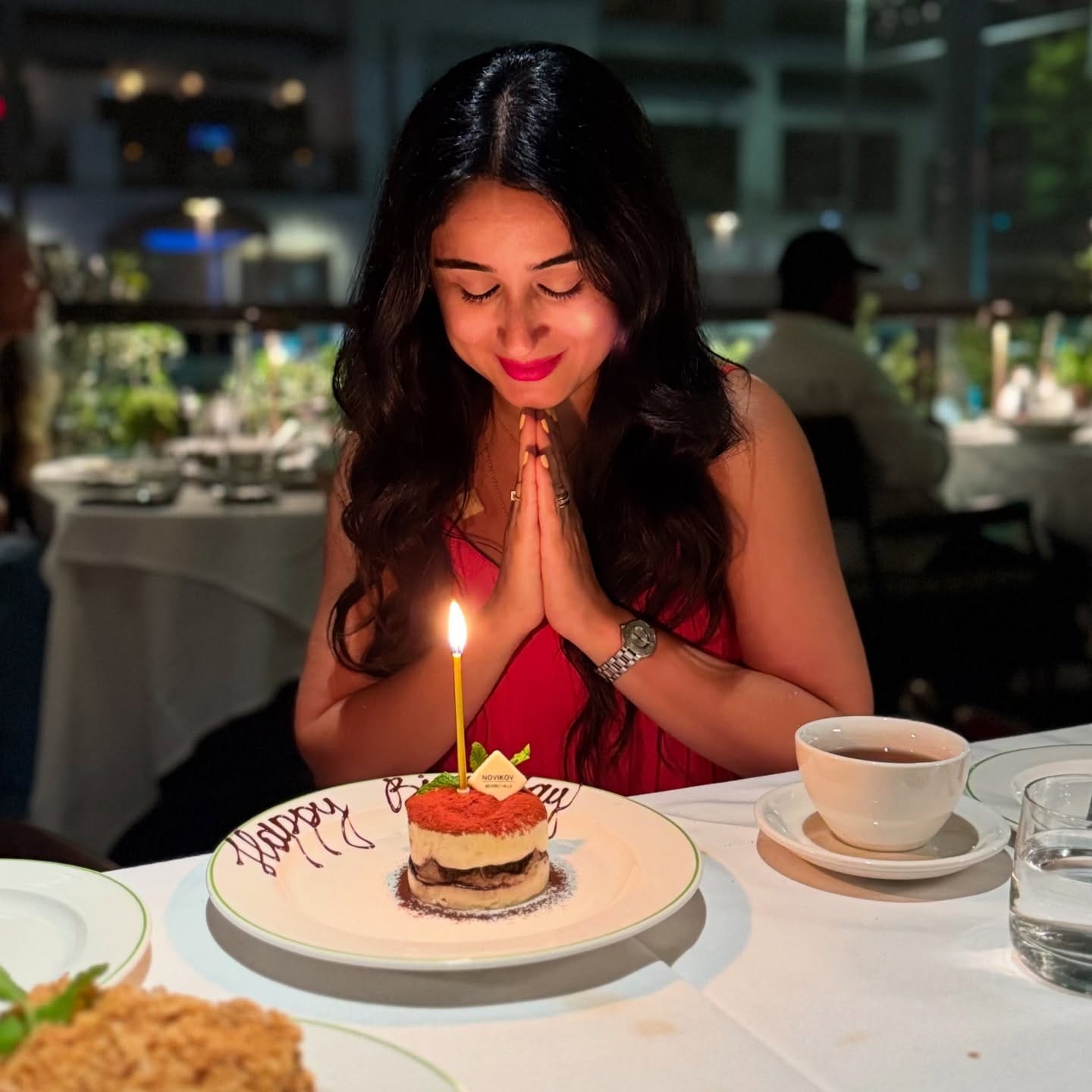 Bita in a red dress with hands pressed together, smiling at a tiramisu with one lit candle on a Happy Birthday plate at a white-clothed restaurant table at night.