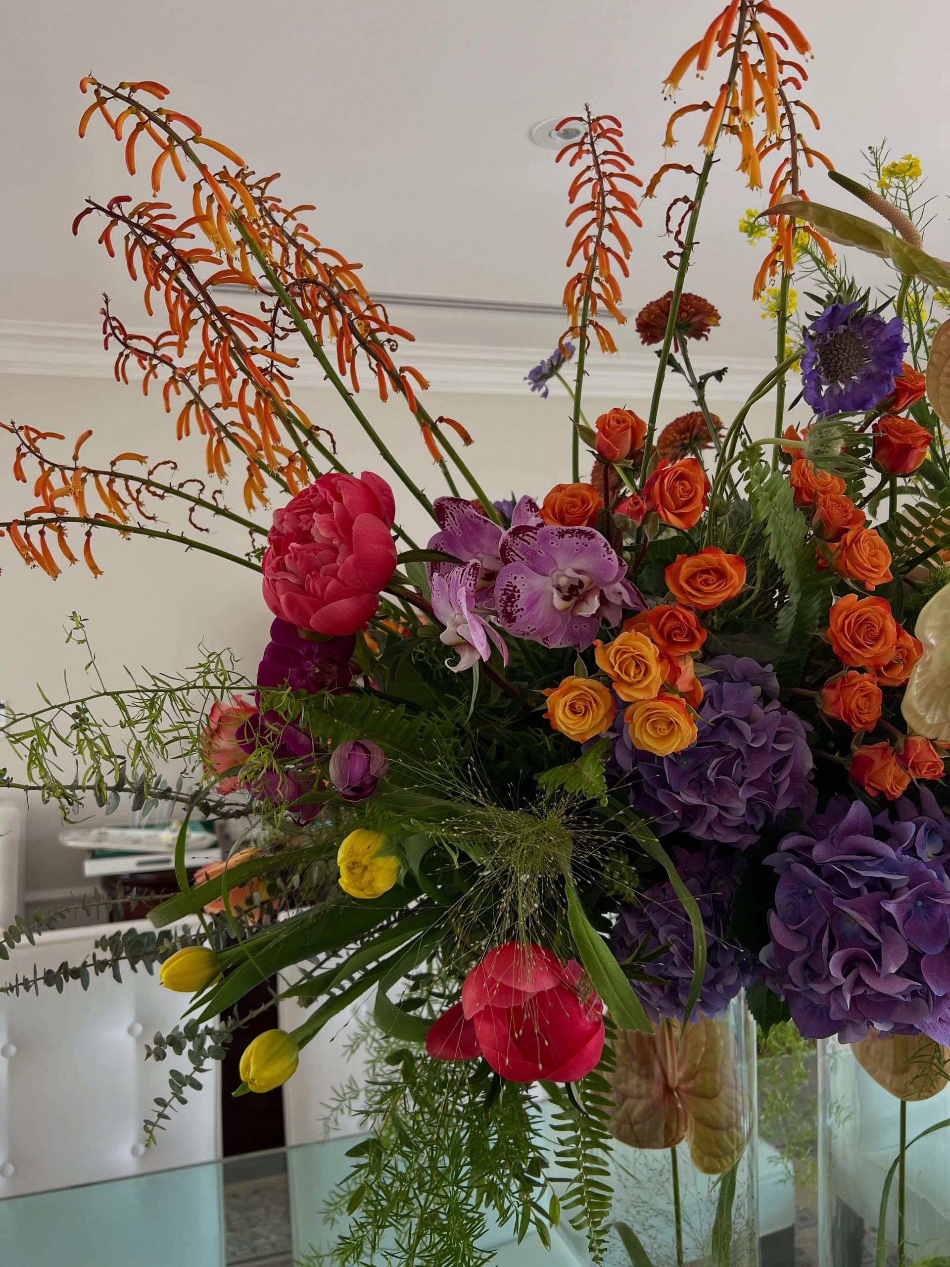 Close-up of orange tubular flower spikes, a coral peony, spotted pink phalaenopsis, orange spray roses, purple hydrangea, purple scabiosa, yellow tulips and eucalyptus.