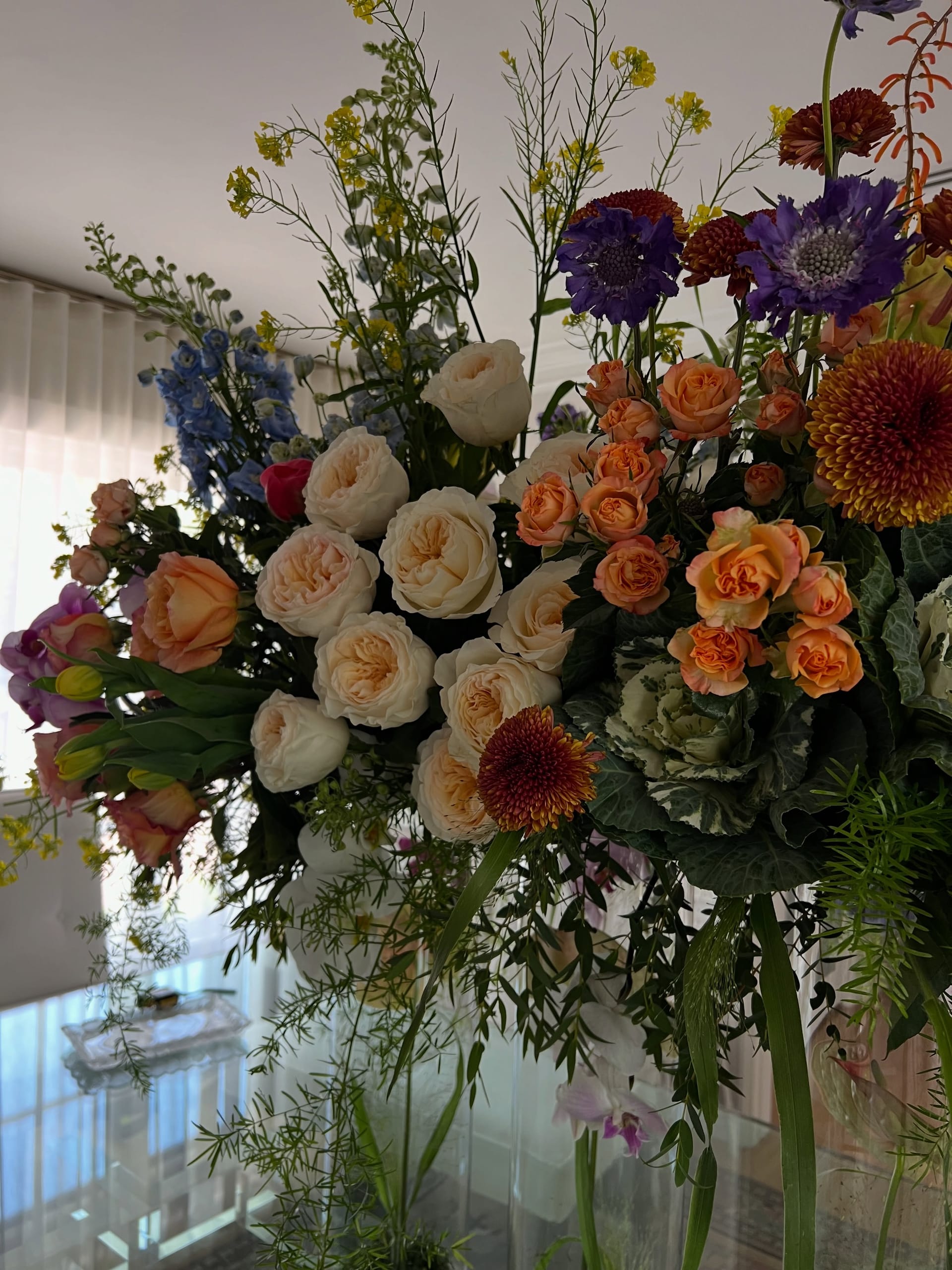 Backlit close-up of cream and peach garden roses, blue delphinium, purple scabiosa, rust chrysanthemums, orange spray roses and ornamental kale against sheer curtains.