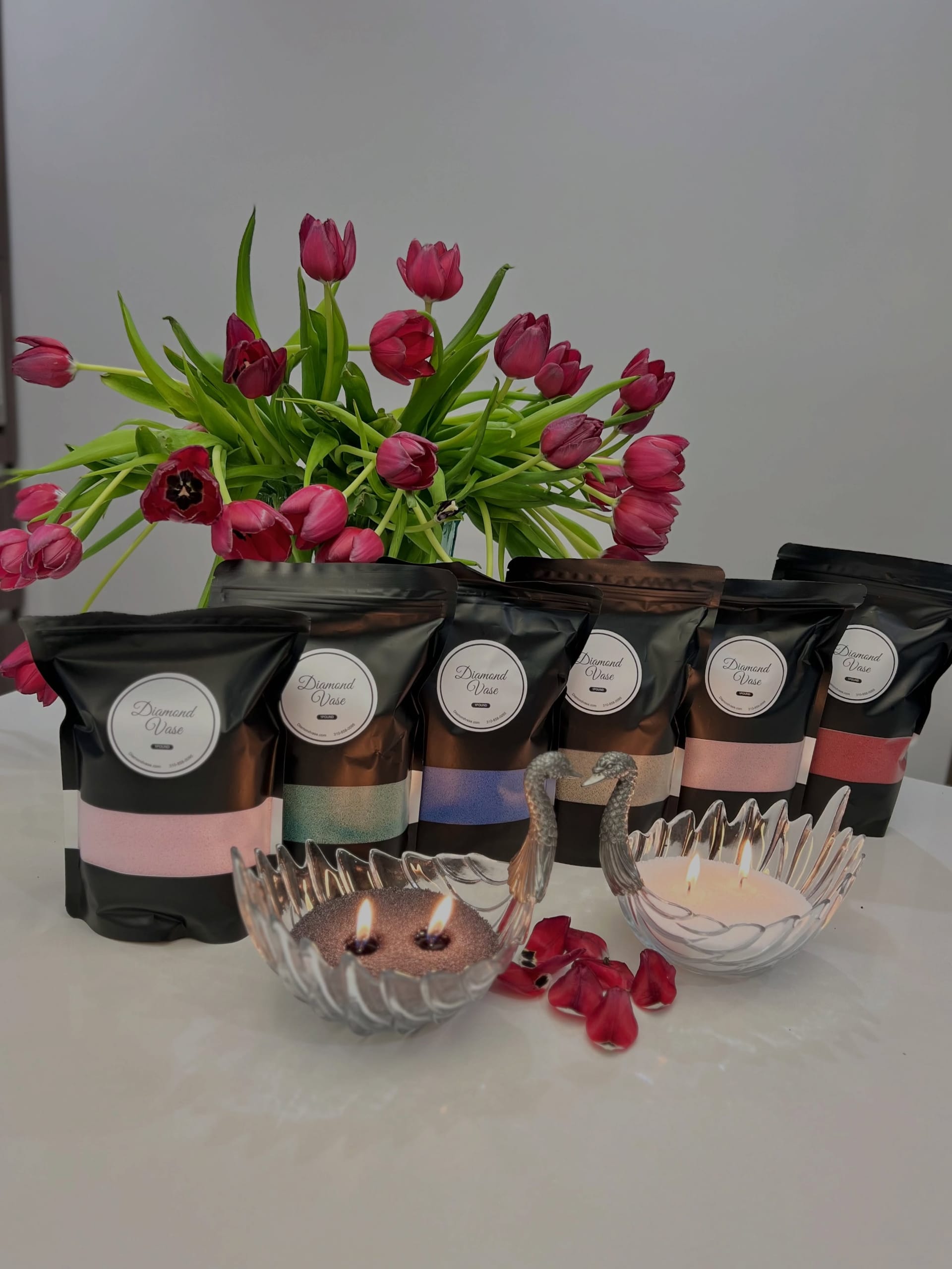 Six black Diamond Vase one-pound pouches of candle sand in pink, teal, blue, gold, pink and red lined up with two glass swan bowls burning pink and white sand candles, magenta tulips and rose petals on a white table.