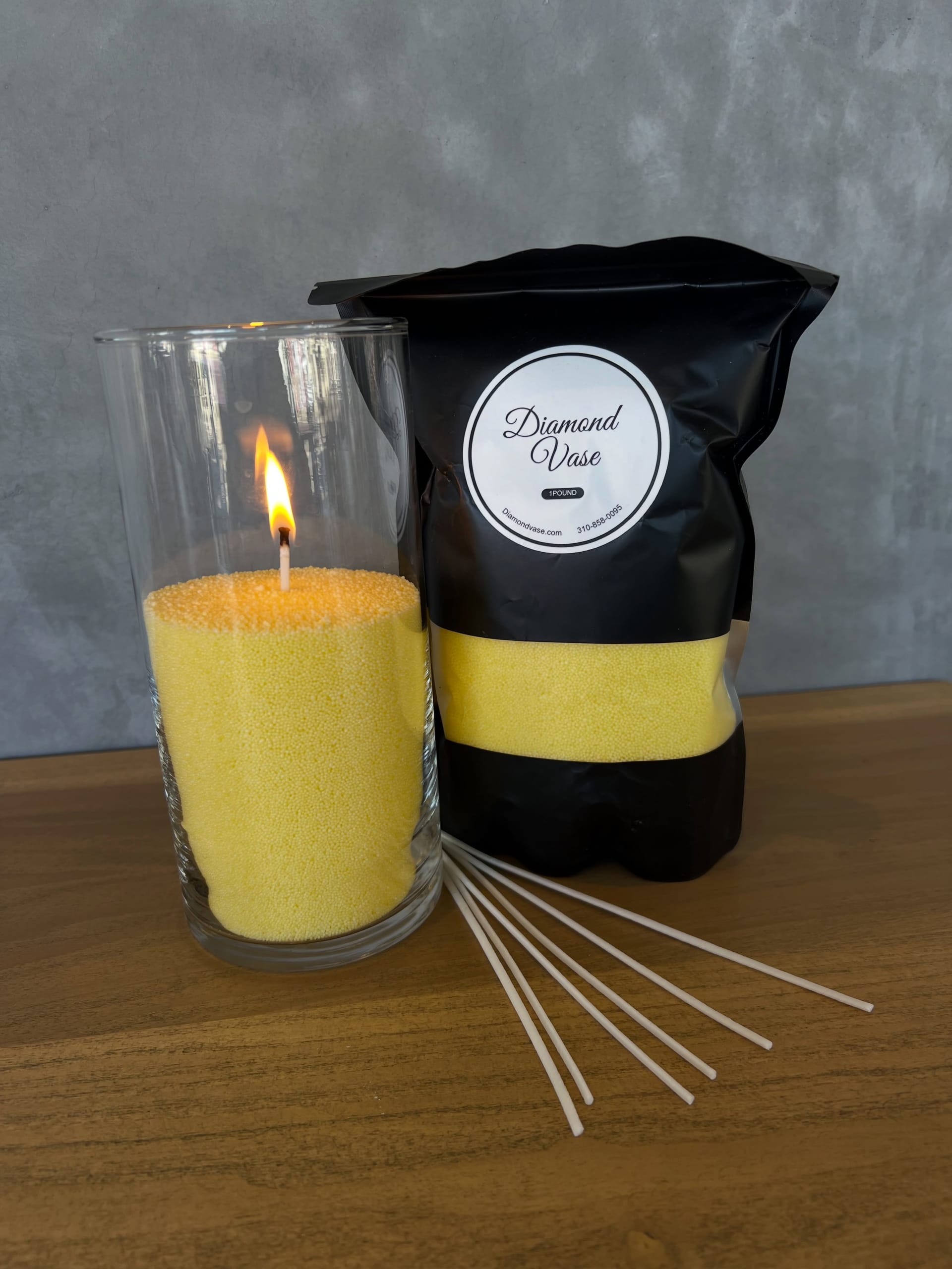 A pale yellow sand candle burning in a clear glass cylinder beside its black Diamond Vase pouch and white wicks on a wood table against a grey concrete wall.