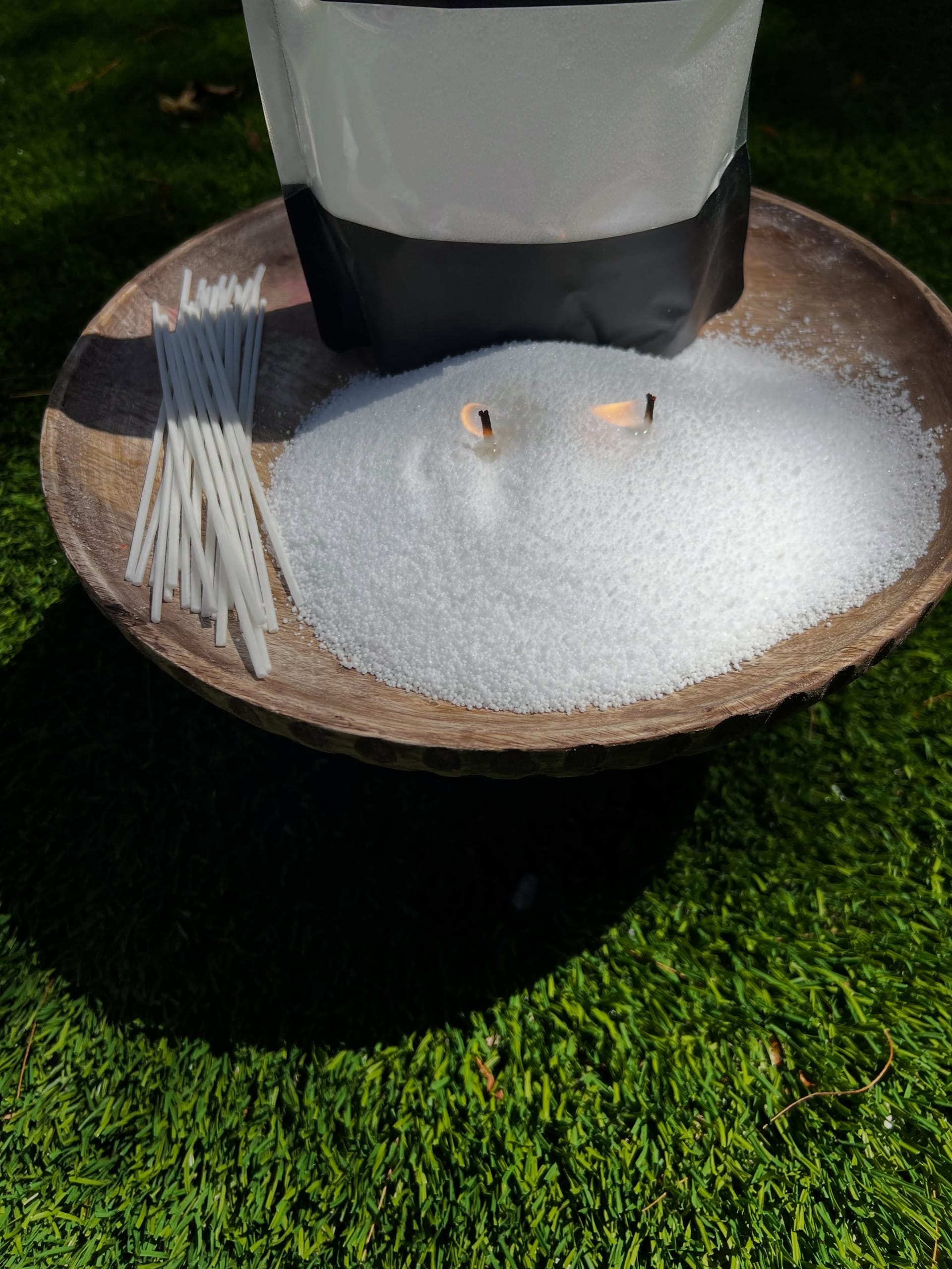 White candle sand with two lit wicks heaped on a wooden pedestal bowl on artificial grass, a bundle of wicks beside it and the pouch cropped at the top.