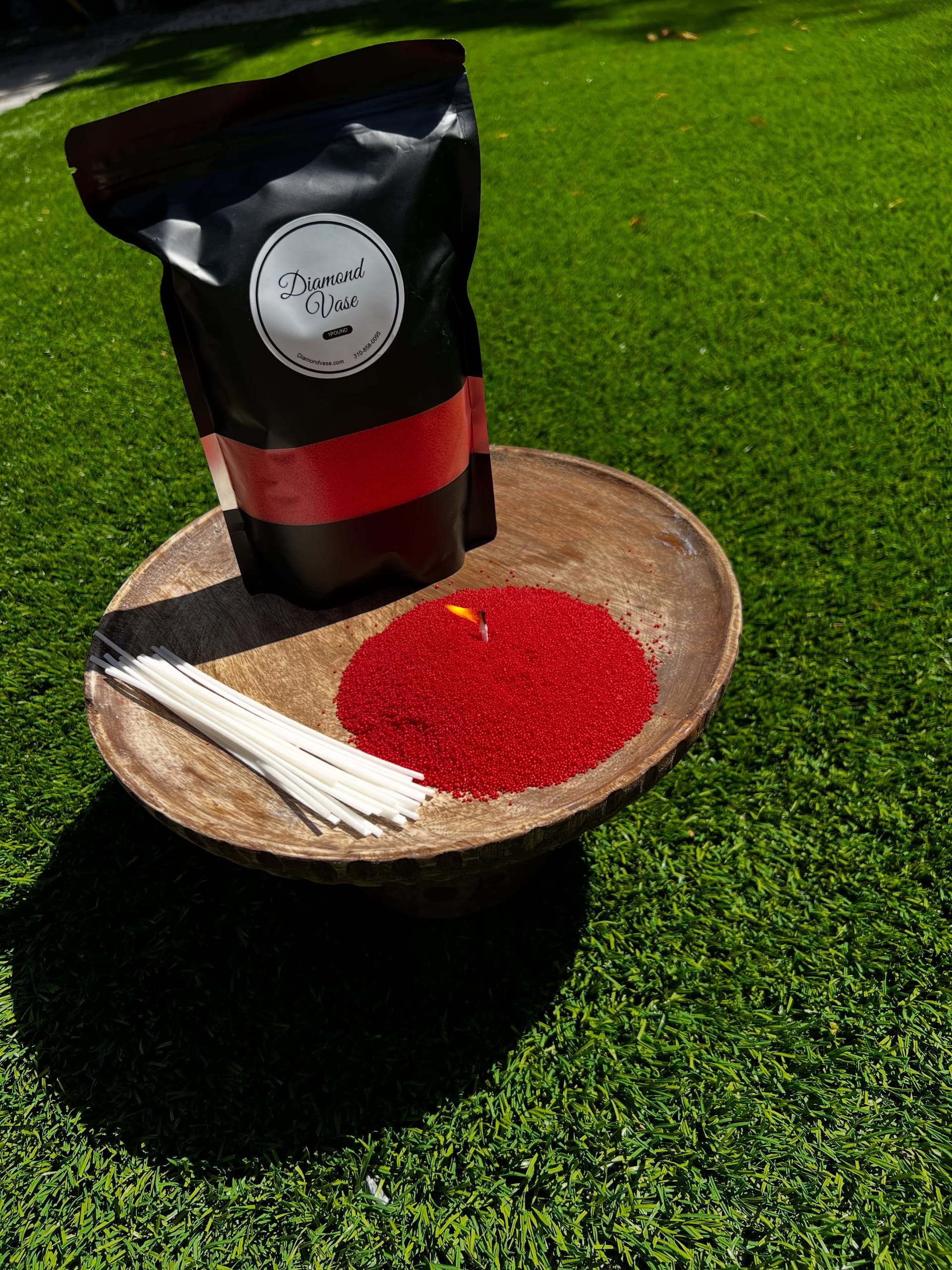 Bright red candle sand with a lit wick on a wooden pedestal bowl on green grass beside a black Diamond Vase pouch with a red window and a bundle of wicks.