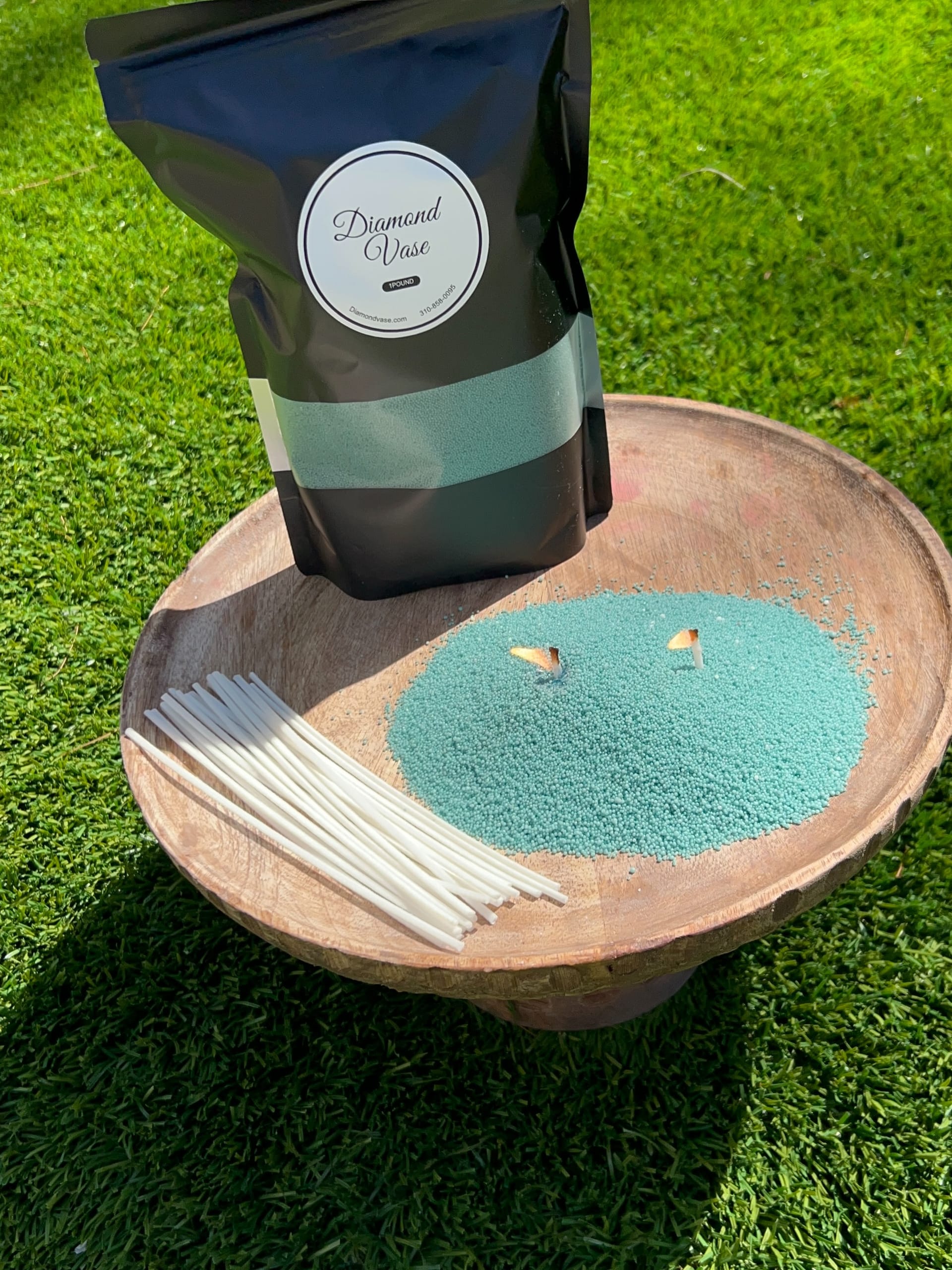 Teal-aqua candle sand with two lit wicks on a wooden pedestal bowl on grass beside a black Diamond Vase pouch with a teal window and white wicks.