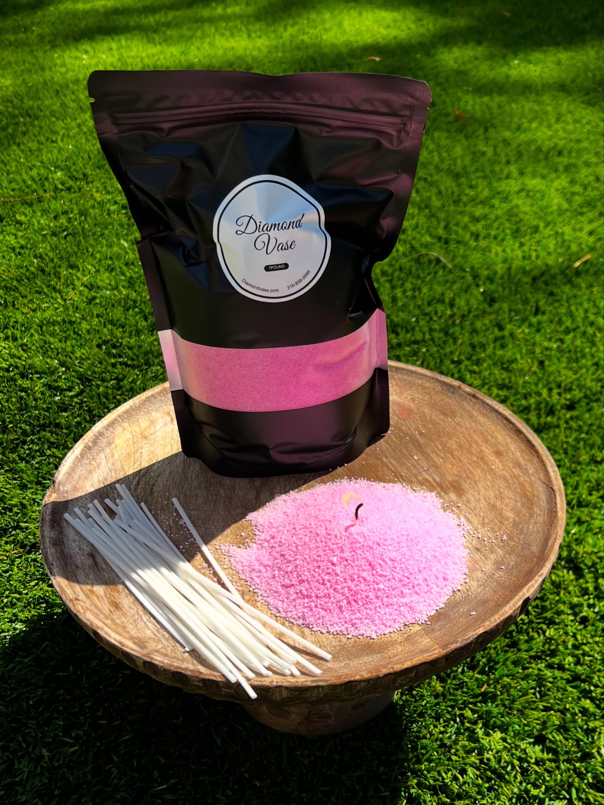 Bubblegum-pink candle sand with a lit wick on a wooden pedestal bowl on grass beside a black Diamond Vase pouch with a pink window and a fan of wicks.